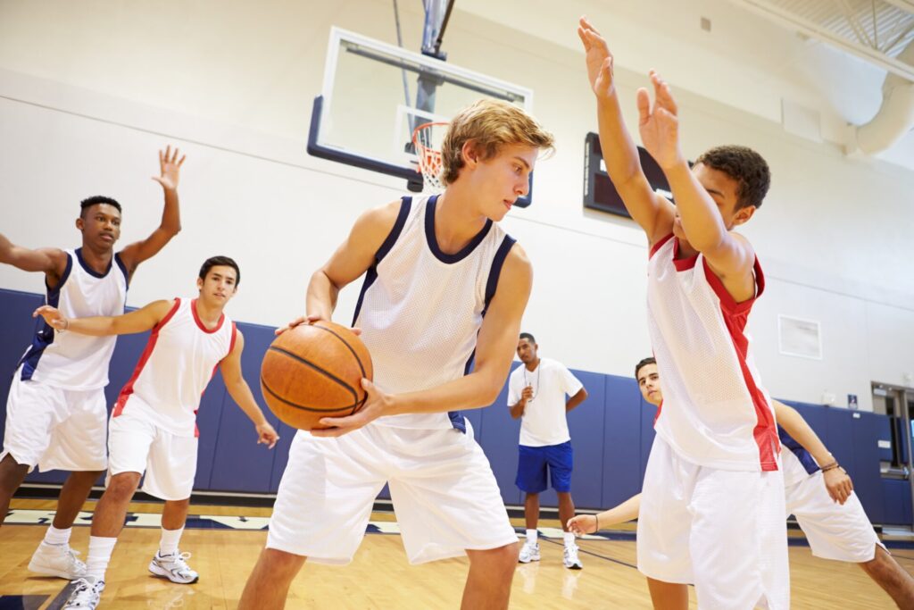 Un joueur de basketball en maillot blanc et bleu tient le ballon et cherche une opportunité de passe tandis qu'un défenseur en maillot blanc et rouge lève les bras pour bloquer son action. D'autres joueurs et un entraîneur sont visibles en arrière-plan dans un gymnase.