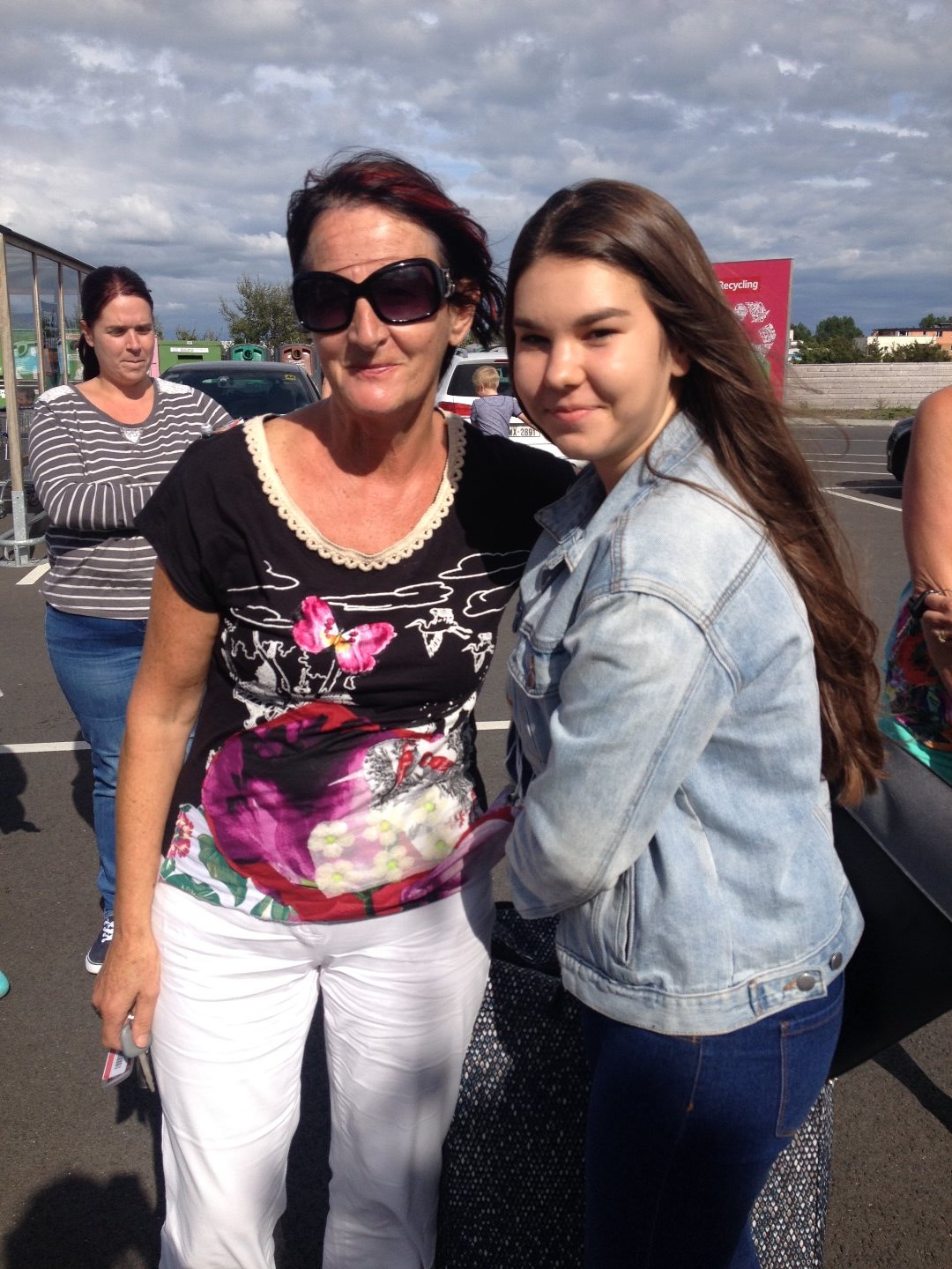 Deux personnes souriantes posent ensemble en extérieur sur un parking sous un ciel partiellement nuageux. Une femme portant des lunettes de soleil et un haut fleuri se tient à côté d'une jeune fille en veste en jean. D'autres personnes sont visibles en arrière-plan.