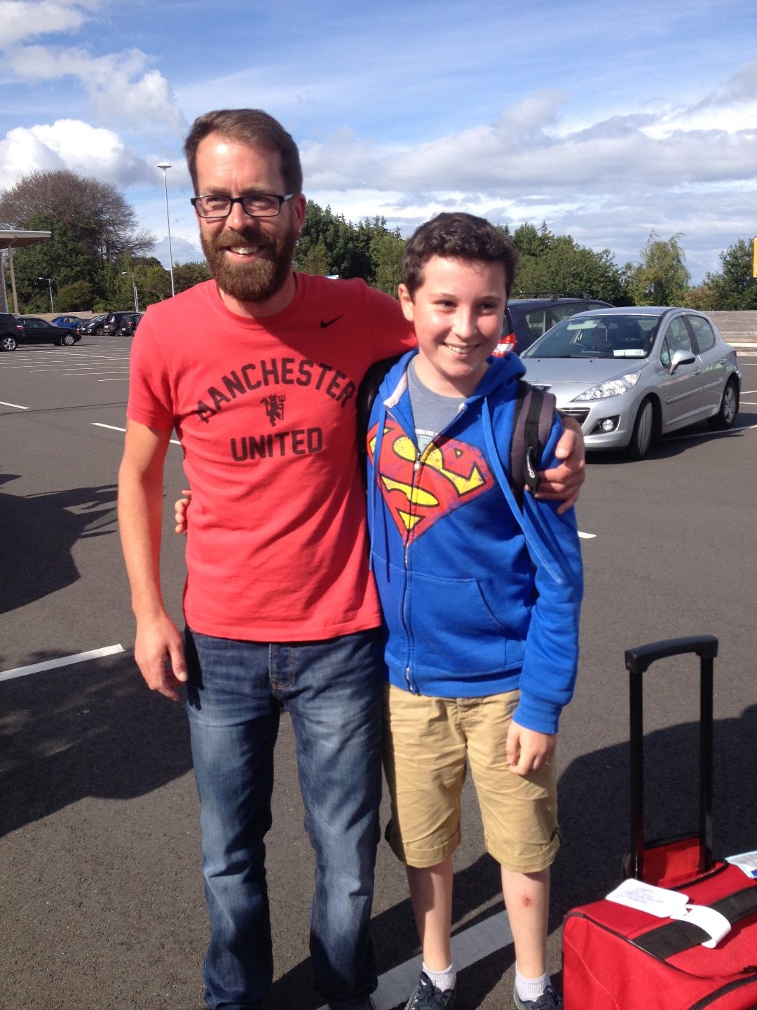 Un homme barbu portant un t-shirt rouge "Manchester United" et un garçon en sweat bleu Superman posent ensemble en souriant, debout sur un parking. À côté d’eux, une valise rouge est posée au sol.