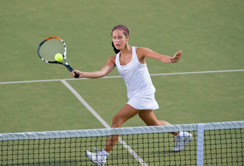 Une jeune joueuse de tennis en tenue blanche effectue une volée sur un court en gazon synthétique, illustrant la concentration et l'intensité d'un stage de tennis en immersion.