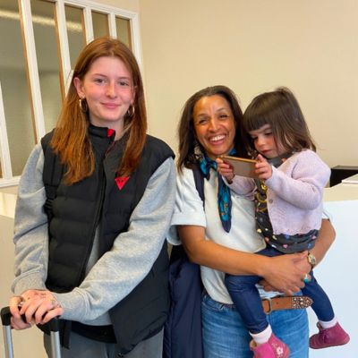Une adolescente souriante, locataire d'une valise, pose aux côtés de sa famille d'accueil à son arrivée en Irlande, sous un ciel partiellement nuageux.